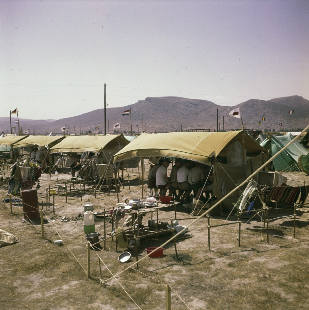 Gruppe von Zelten auf einem grünen Feld mit Menschen, die darauf sitzen und stehen, Fahnen auf Stangen im Hintergrund, Hügel unter einem klaren blauen Himmel und verstreute Campinggegenstände.