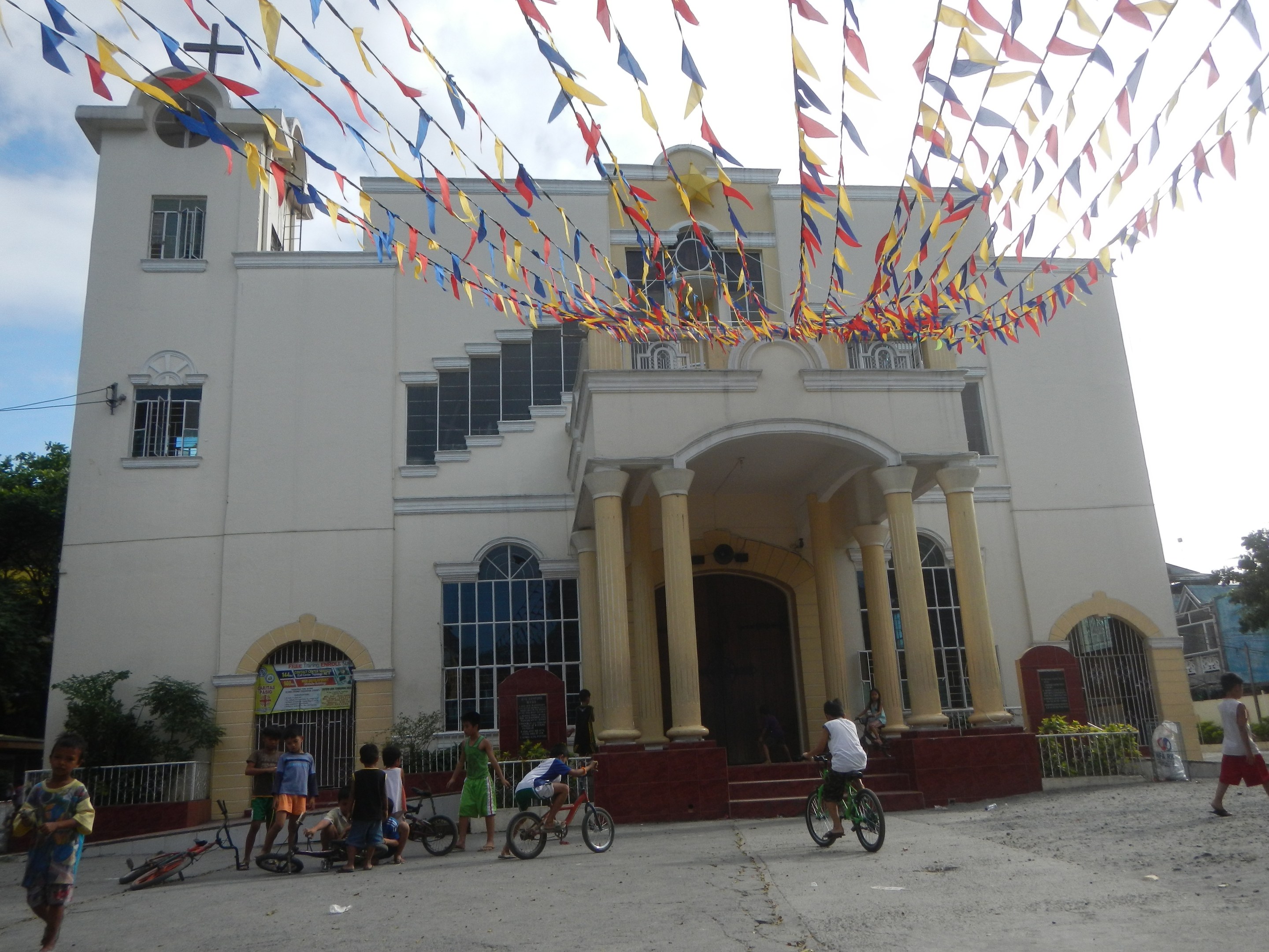 Eine Gruppe von Menschen auf Fahrrädern vor einer weißen Kirche mit architektonischen Details, Pflanzen, einem Zaun, einer Fahne, Bäumen und einem bewölkten Himmel, mit bunten Fahnen an einem Seil oben.