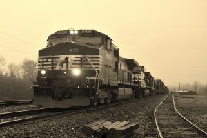 Ein Zug auf Schienen mit Autos, Strommasten, Bäumen und einem Himmel im Hintergrund.