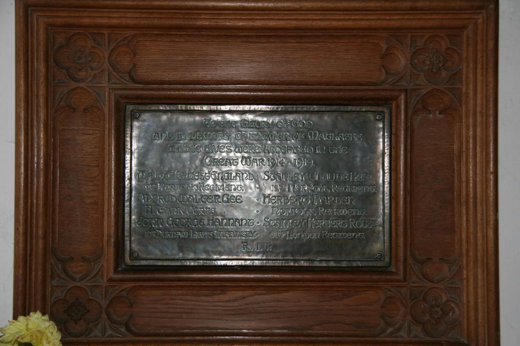 Eine Gedenktafel mit Text ist an einer braunen Holztür oder Wand angebracht, mit einer gelben Blumenvase in der unteren linken Ecke.