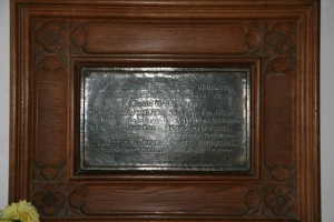 Eine Gedenktafel mit Text ist an einer braunen Holztür oder Wand angebracht, mit einer gelben Blumenvase in der unteren linken Ecke.