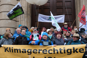 Eine Gruppe von Menschen vor einem Gebäude mit Säulen und einer Tür, die eine Fahne mit Text, Fahnenstangen und Luftballons halten, nehmen an einem Klimastreik in Deutschland teil.