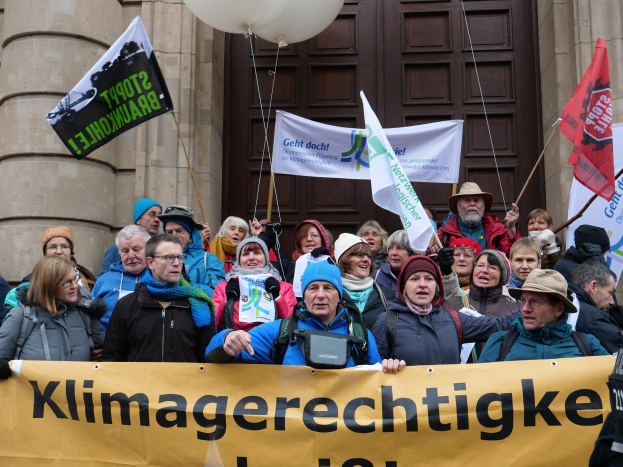 Eine Gruppe von Menschen vor einem Gebäude mit Säulen und einer Tür, die eine Fahne mit Text, Fahnenstangen und Luftballons halten, nehmen an einem Klimastreik in Deutschland teil.