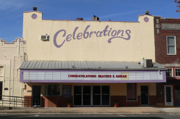 Theatergebäude mit "Feierlichkeiten"-Schriftzug, umgeben von städtischen Strukturen, einer Straße, einem Baum und einem Himmel-Hintergrund.