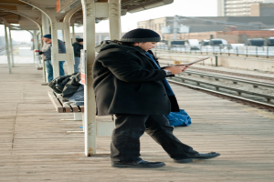 Ein Mann in einer schwarzen Jacke steht an einem Bahnhof, mit einem Eisenstab und einer Hütte dahinter und Bahnschienen und braunen Gebäuden rechts.