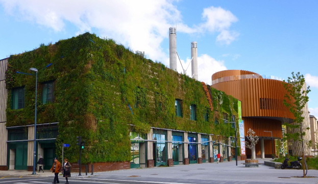 Ein großes Gebäude mit einer grünen Wand an seiner Seite, umgeben von Straßenmasten, Straßenlaternen, Verkehrspfosten, Verkehrssignalen, Kraftfahrzeugen, Fußgängern, Bäumen und einem bewölkten Himmel, als Teil des Green Building Project.