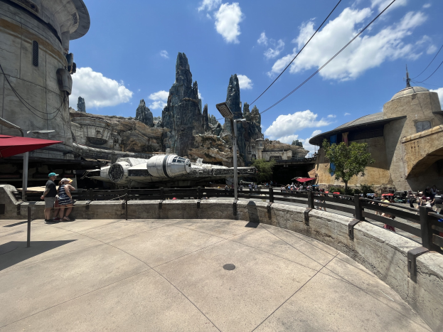 Das Millennium Falken in Star Wars: Galaxy's Edge, umgeben von Menschen mit Schirmen, einem Zaun, einer Straßenlaterne, einem Schild, Bäumen, Hügeln, Drähten und einem bewölkten Himmel.
