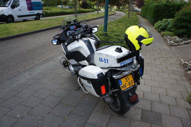 Ein polizeiliches Motorrad am Straßenrand mit einem Helm darauf, mit Bäumen, Pfählen, Gebäuden, Pflanzen, Gras, Steinen und anderen Fahrzeugen im Hintergrund.
