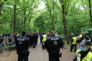 Eine Gruppe von Polizisten steht vor einer Menge von Menschen, einige tragen Mützen und Masken, mit Fahrrädern und einer Bank im Vordergrund und Bäumen und Himmel im Hintergrund, während einer Anti-Terror-Demonstration in Berlin.