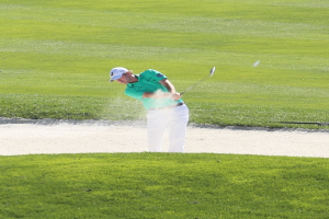 Ein Mann in einem weißen Hemd und schwarzen Hosen schwingt einen Golfschläger auf einem Kurs mit grünem Gras, einem Wasserhindernis und mehr Gras im Hintergrund.