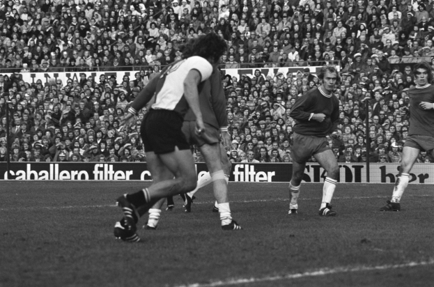 Schwarzes und weißes Foto von Männern in Sportuniformen, die Fußball auf einem Feld mit einer großen Menge im Hintergrund spielen, Tafeln mit Text, Geländer und Pfosten sichtbar.