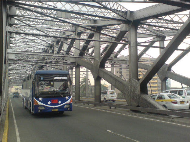 Ein blauer Bus fährt über eine Brücke mit Autos, umgeben von Gebäuden, Bäumen, Pfählen und einem klaren blauen Himmel.