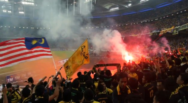 Eine große Menschenmenge in einem Stadion mit brennenden Fackeln in der Luft, einige halten Fahnen und eine Person hält eine Kamera, mit Rauch im Hintergrund und Tafeln mit der Aufschrift "Malaysia vs Malaysia Live Streaming".