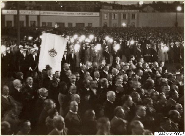 Schwarzes und weißes Foto einer großen Menschenmenge vor einem Gebäude, mit einer Person, die eine Fahne hält, in der Mitte, Polen und Lichter im Hintergrund und Text, der unten 'Gustav Klimts Rede an der Berliner Mauer' lautet.