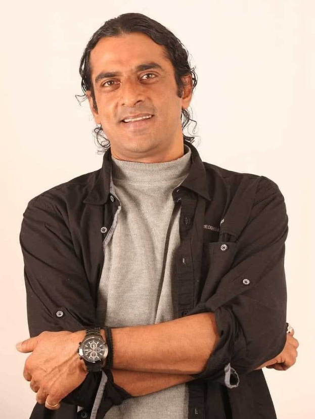 Ein Mann in einem schwarzen und aschfarbenen Kleid mit verschränkten Armen, der eine Uhr trägt und vor einem weißen Hintergrund steht und nachdenklich wirkt.