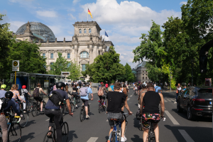 Eine Gruppe von Menschen, die auf Fahrrädern eine von Bäumen gesäumte Straße in Berlin, Deutschland, entlangfährt, mit Gebäuden, einer Bushaltestelle auf der rechten Seite und einer Flagge, die auf einem der Gebäude weht, unter einem bewölkten Himmel.