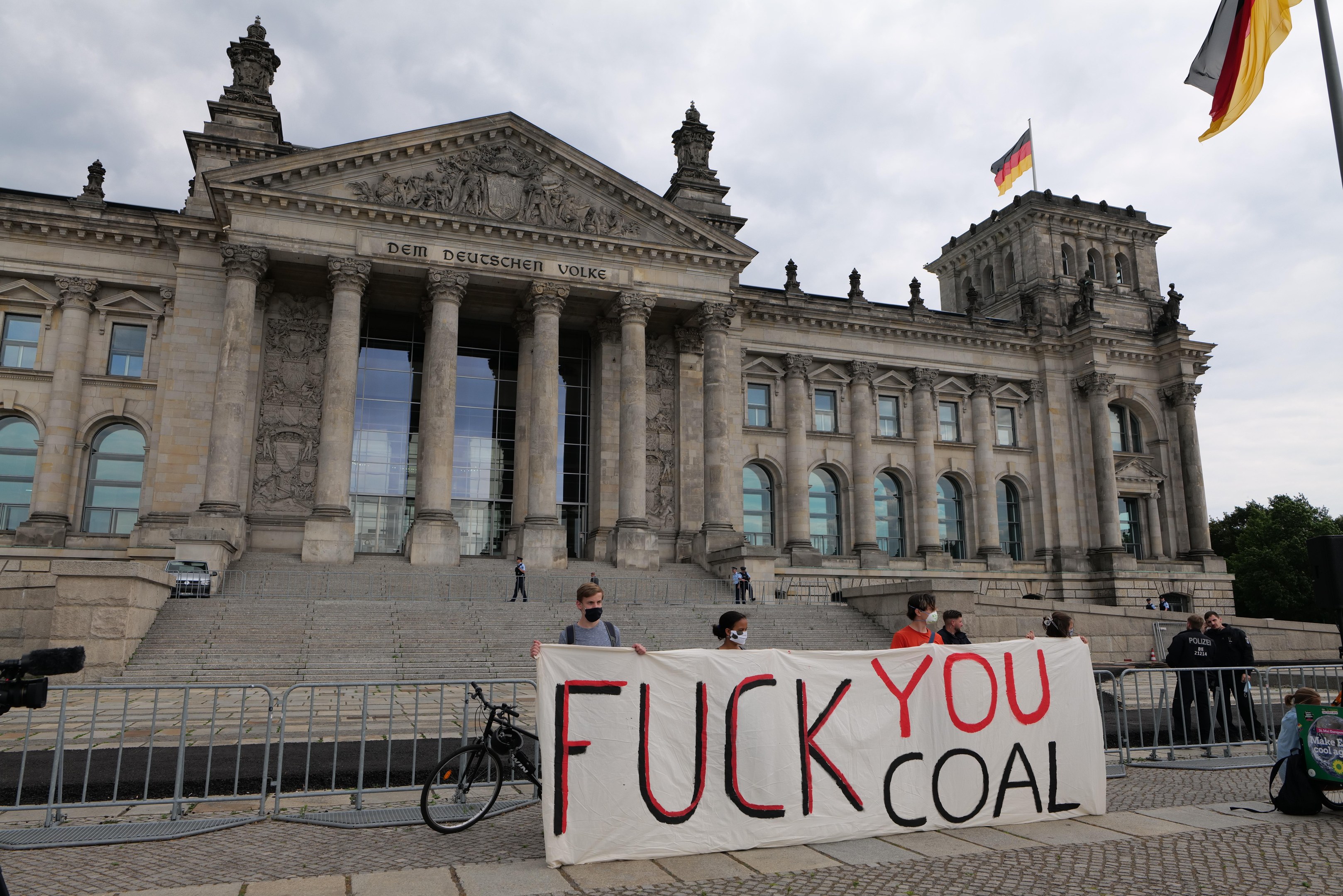 Eine Gruppe von Menschen hält ein Transparent mit der Aufschrift "Fuck You Coal" vor dem Reichstagsgebäude in Berlin, Deutschland, mit Bäumen und einer Fahnenstange im Hintergrund.