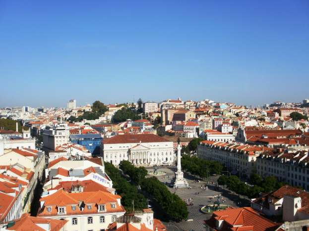 Blick auf Lissabon von einem Hügel mit Gebäuden mit Fenstern, Bäumen, einer Statue auf einem Sockel, einer Straße mit wenigen Menschen und dem Himmel im Hintergrund.