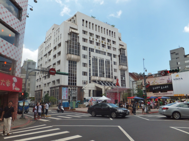 Eine belebte Stadtstraße mit Fußgängern, die vor einem hohen Gebäude über die Straße gehen, Fahrzeuge auf der Straße und Verkehrsampeln neben Gehwegen, Bäumen und Gebäuden im Hintergrund.