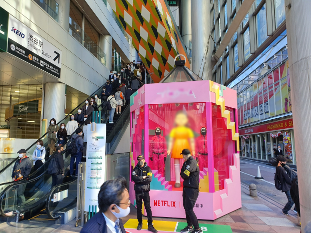 Eine Gruppe von Menschen steht vor einem Gebäude mit Glaswänden, Säulen und Texttafeln, einige tragen Masken, während eine Rolltreppe mit Menschen in der Nähe nach oben führt; eine pinkfarbene Box in der Mitte zeigt Schaufensterpuppen und trägt den Text "Netflix's new Netflix booth in Tokyo."