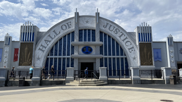 Außenansicht des Eingangs zum Hall of Justice im Disney's Hollywood Studios mit einer Gruppe von Menschen davor, Bannern am Gebäude und einer bewölkten Himmel.