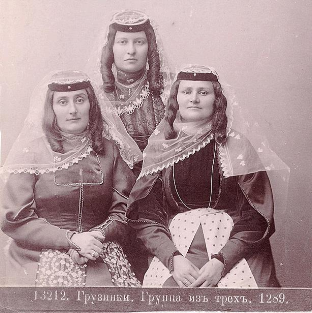 Drei Frauen in traditionellen russischen Hochzeitskleidern mit aufwendiger Stickerei und Perlen stehen vor einer weißen Wand, mit Text unten, der angibt, dass es ein russisches Hochzeitsfoto ist.