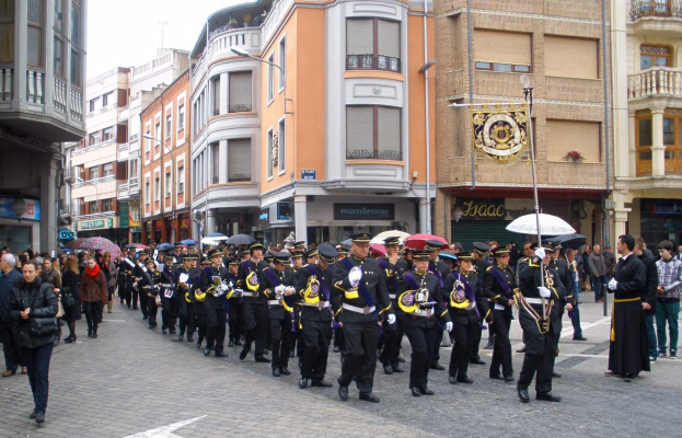 Eine Gruppe von Menschen, die mit Schirmen auf der Straße gehen, einige halten Musikinstrumente und andere tragen Taschen, mit Gebäuden, Laternenmasten und Namensschildern im Hintergrund unter einem sichtbaren Himmel, während eines Ehrenzuges.