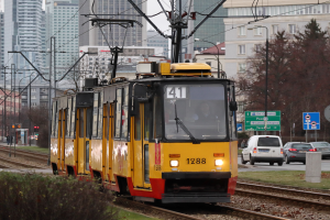 Eine gelbe und rote Straßenbahn fährt auf Schienen neben hohen Gebäuden, mit Strommasten, Drähten, Fahrzeugen, Schildern, Bäumen und einem klaren blauen Himmel.