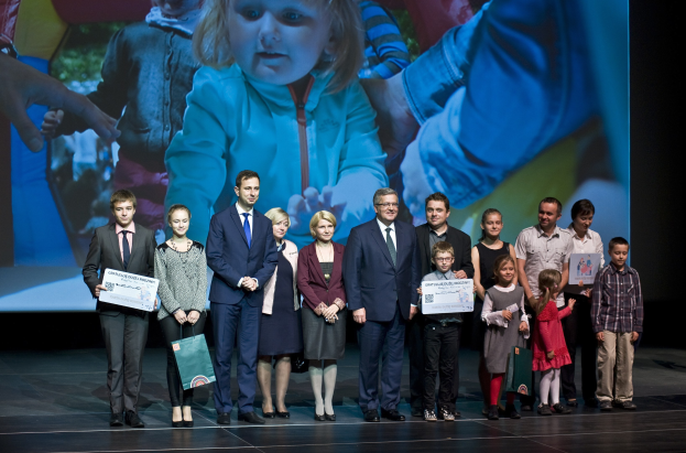 Eine Gruppe von Menschen auf einer Bühne mit Taschen und Urkunden, im Hintergrund ein Bildschirm mit dem Bild eines Kindes und anderen Personen.
