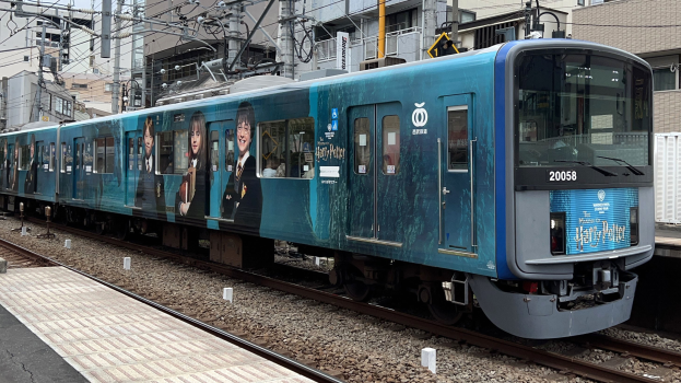 Blauer Zug fährt auf Schienen neben einem Bahnsteig mit Strommasten und Drähten, Gebäuden und einem klaren Himmel im Hintergrund.