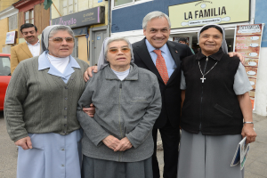 Eine Gruppe von Menschen, die lächeln und stehen, mit einer Frau rechts, die eine Zeitung hält, vor einem Gebäude mit einer Wand, einer Tür, Bannern, einem Auto und einer weiteren Person im Hintergrund.