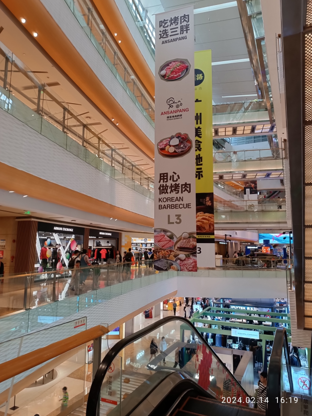Ein Innenblick in ein Einkaufszentrum mit einer Rolltreppe in der Mitte, Menschen mit Handtaschen, von der Decke hängende Banner mit Text und Läden im Hintergrund.