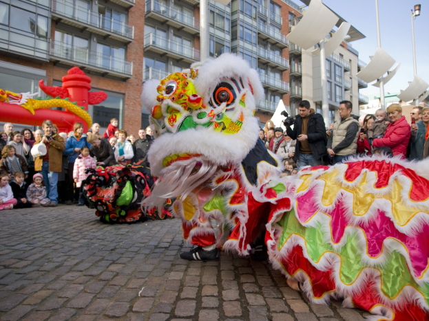 Ein lebendiges chinesisches Neujahrsfest in Amsterdam mit einem Löwen tanzen im Vordergrund und einer Menschenmenge drumherum, einige halten Kameras, mit Gebäuden, Laternenmasten und einem klaren blauen Himmel im Hintergrund.