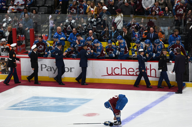 Ein Eishockeyspieler in einer Spielsituation umgeben von Teamkollegen und Zuschauern, mit Werbetafeln im Hintergrund, die ein Duell zwischen Colorado Avalanche und San Jose Sharks zeigen.