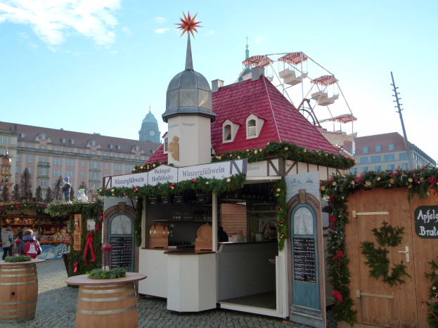 Ein kleines Gebäude mit einem roten Dach und einem Riesenrad davor, umgeben von Menschen und Festdekorationen bei einem Outdoor-Event in München, Deutschland.