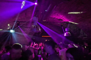 Eine Gruppe von Menschen tanzt in einem Nachtclub unter lila und blauen Deckenlichtern, einige halten Flaschen.