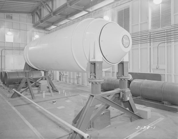 Ein Schwarz-Weiß-Foto einer großen zylindrischen Wasserstoffbombe auf einem Ständer in einer industriellen Fabrikumgebung mit Rohren, Beleuchtung und einer Wand im Hintergrund.