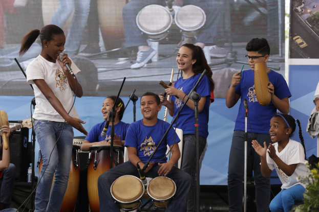 Gruppe von Kindern, die Musikinstrumente auf einer Bühne spielen, mit einer Frau, die ein Mikrofon hält, in der Mitte, umgeben von Mikrofonstativen, einem Banner im Hintergrund und Pflanzen auf der rechten Seite.