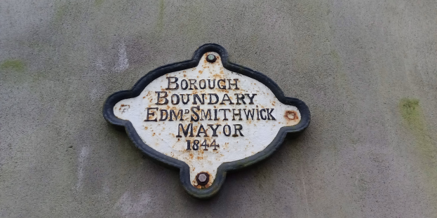 Schwarze Metallplatte mit weißer, fetter, zentrierter Schrift, die "Borough Boundary Edm Smithwick Mayor" auf der Seite eines Gebäudes liest.