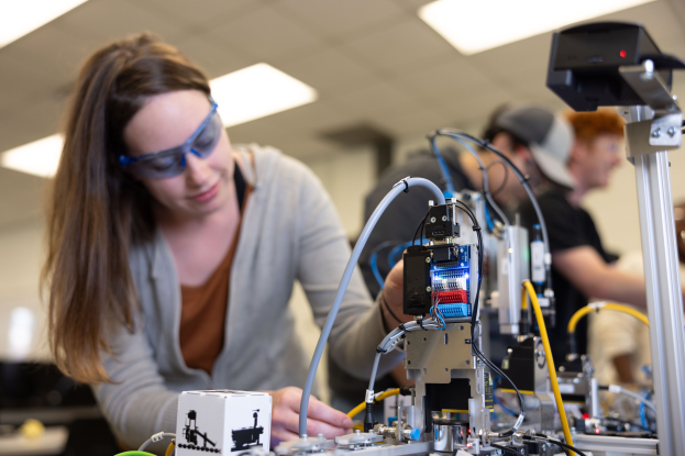 Eine Frau in Schutzbrille arbeitet an einem Roboter in einem Labor umgeben von anderen, mit Werkzeugen und Komponenten auf dem Tisch.