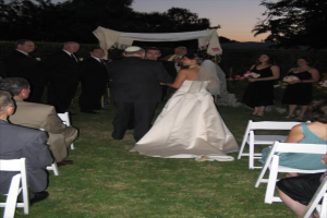 Eine Gruppe von Menschen bei einer Hochzeit, wobei Frauen rechts Blumensträuße halten, während andere auf Stühlen sitzen und Bäume im Hintergrund zu sehen sind.