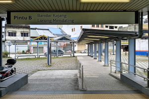 Ein Motorrad steht vor einem Gebäude mit der Aufschrift "Pintu B Bandar Kajang Lorong Mendaling", mit Fahrzeugen auf der Straße, Gebäuden mit Fenstern, einem Schuppen mit Säulen, Geländern, Deckenleuchten, Bäumen und einem klaren blauen Himmel im Hintergrund.