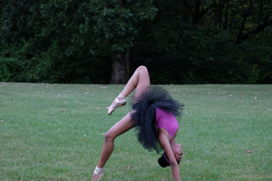 Eine Frau in einem pinken und schwarzen Kleid führt Gymnastik auf Gras mit ein paar Bäumen im Hintergrund aus.