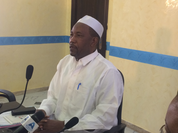 Ein Mann in einem weißen Gewand und Hut sitzt an einem Tisch mit Mikrofonen, spricht, während Papiere und Gegenstände auf dem Tisch sichtbar sind, mit einem anderen Menschen mit Brille rechts von ihm, in einem Raum mit einer Wand und einer Tür im Hintergrund.