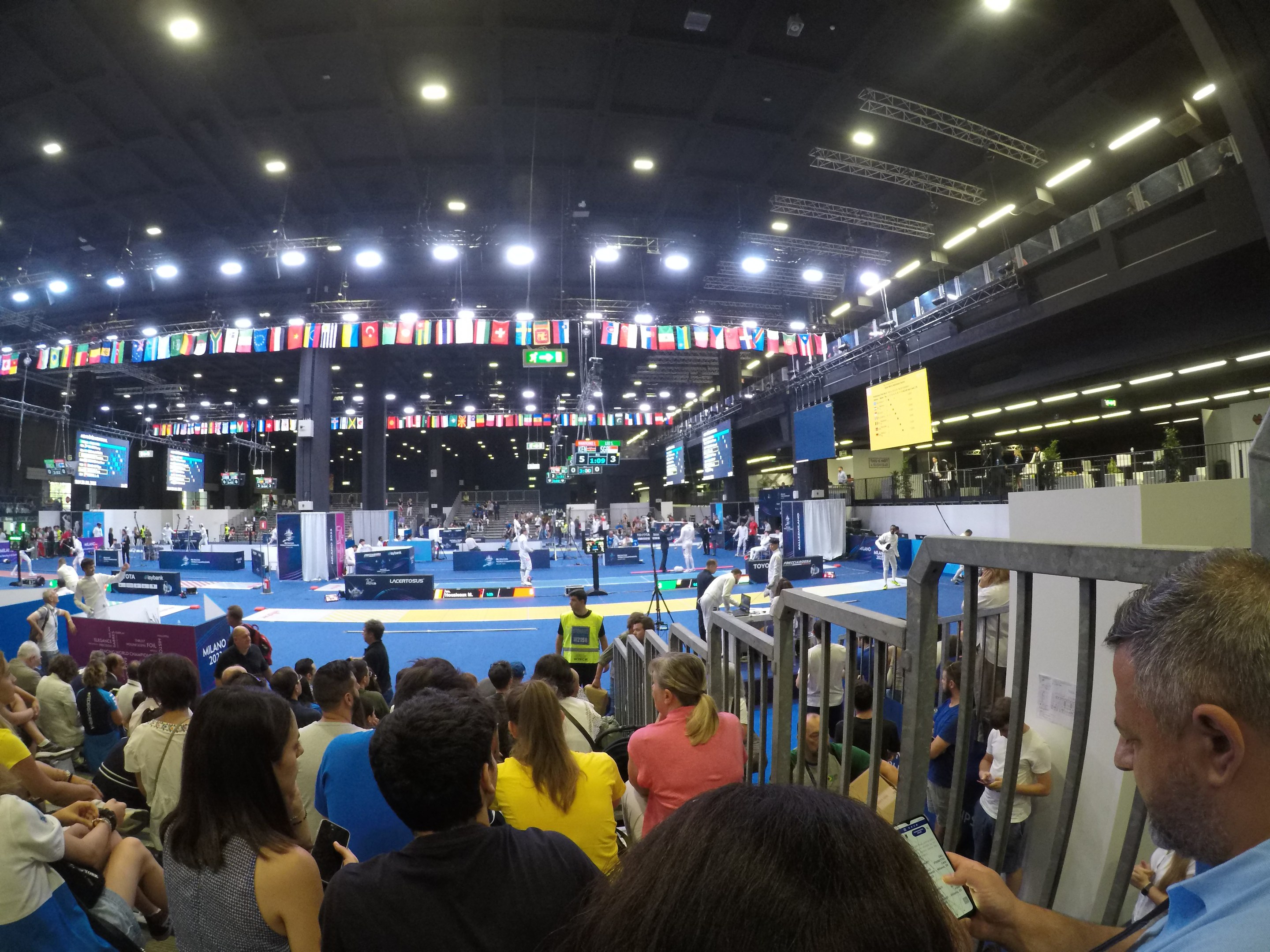 Große Menschenmenge sitzend und stehend in einer Indoor-Sportarena mit Bildschirmen, Flaggen und Deckenbeleuchtung.