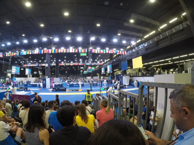 Große Menschenmenge sitzend und stehend in einer Indoor-Sportarena mit Bildschirmen, Flaggen und Deckenbeleuchtung.