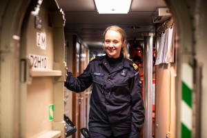 Eine Frau in schwedischer Polizeiuniform lächelt in einem Flur mit nummerierten Türen auf der linken Seite und Papieren auf der rechten Seite, unter einer Deckenlampe.