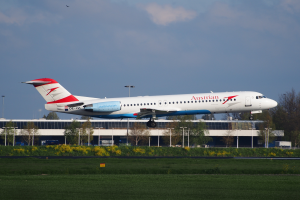 Ein Airbus A320-200 der Austrian Airlines beim Start am Frankfurter Flughafen, mit grünem Gras, blühenden Pflanzen, Bäumen, Laternenmasten und einem Gebäude im Hintergrund bei bewölktem Himmel.