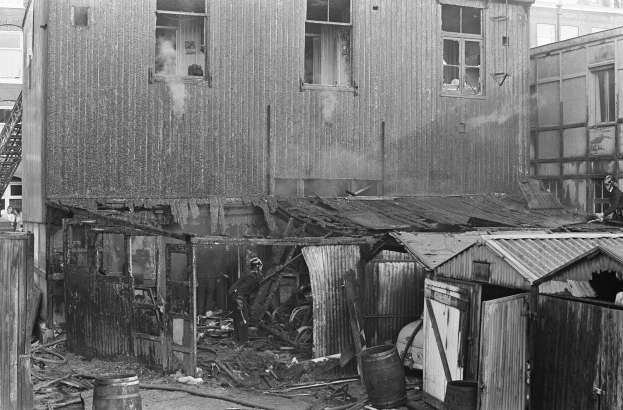 Ein Schwarz-Weiß-Foto eines durch Feuer beschädigten Gebäudes mit verstreuten Trümmern, darunter Fässer und hölzerne Gegenstände, und einigen Menschen in der Nähe, mit anderen Gebäuden, einer Leiter und einem Pfahl im Hintergrund.