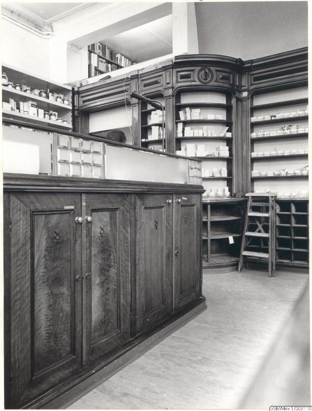 Schwarzes und weißes Foto eines vintage Apothekeninneren mit einer Theke, Schränken, einer Leiter und Regalen, die mit verschiedenen Gegenständen gefüllt sind, mit Text oben.